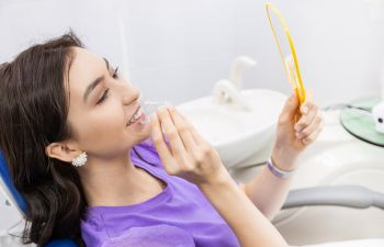 A satisfied woman patient takin off clear orthodontic braces and watching her teeth in a mirror.