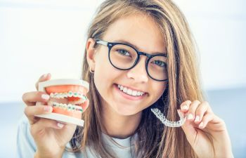 A smiling teenage girl holding an invisible orthodontic aligner in one hand and a teeth model with traditional braces in the other one.